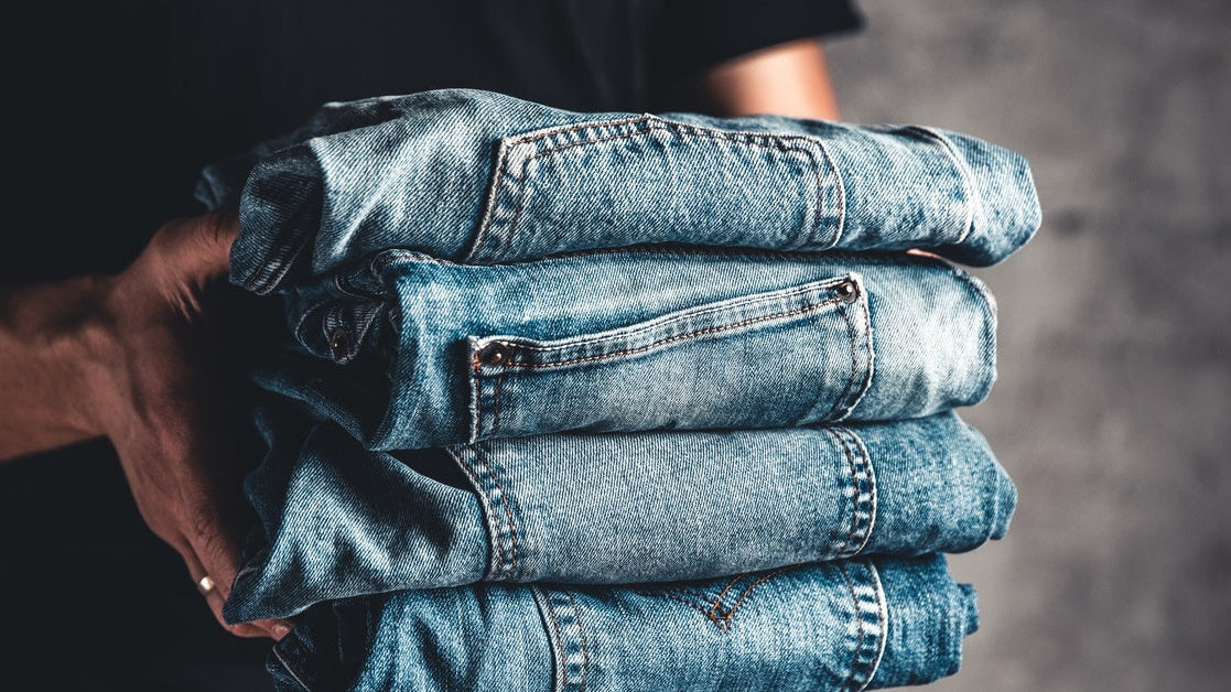 A man holding a stack of four folded blue jeans, featuring slightly different colors and pocket designs.