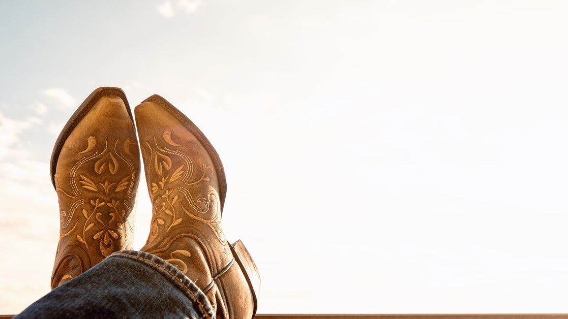 Someone wearing a pair of cowboy boots and propping their feet up, ankles crossed, on a wooden beam.