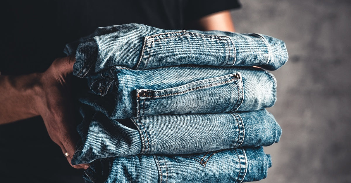 A man holding a stack of four folded blue jeans, featuring slightly different colors and pocket designs.
