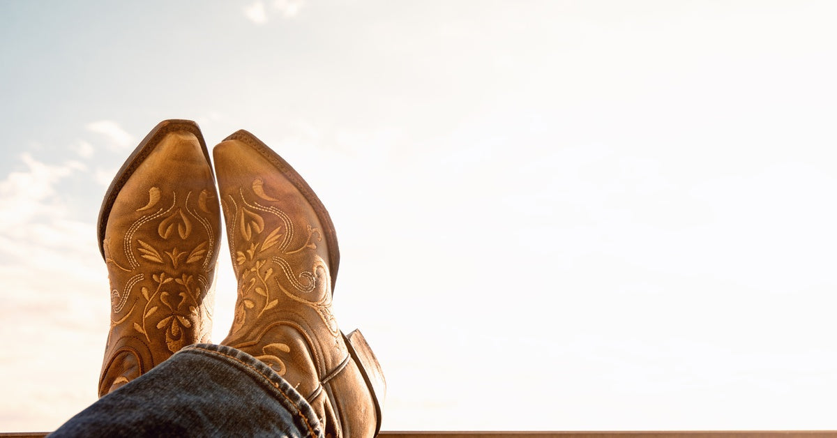 Someone wearing a pair of cowboy boots and propping their feet up, ankles crossed, on a wooden beam.